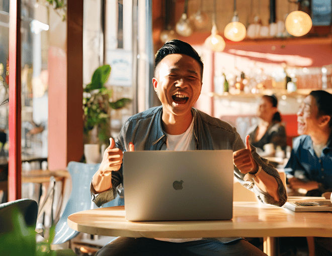 Sunlight bathes the café, illuminating an Indonesian man who clenches his fist in excitement at his laptop. The atmosphere is vibrant with modern decor and patrons.